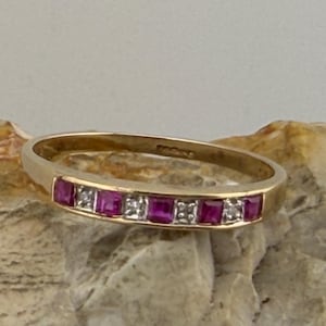 Könnte beinhalten: Ein goldener Bandring mit abwechselnd quadratisch geschliffenen Rubinen und kleinen Diamanten. Die Edelsteine des Rings sind leuchtend rot und weiß. Der Ring wird vor einem neutralen Hintergrund präsentiert. Ein klassisches Schmuckstück.