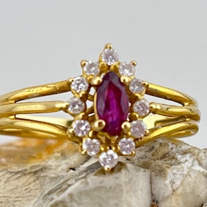May include: A gold ring with a red ruby gemstone surrounded by a halo of smaller clear gemstones. The ring has a triple band design.