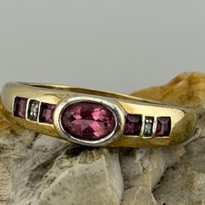 May include: A gold ring with a central oval-cut, deep pink gemstone, flanked by smaller square gemstones and accent diamonds. The ring has a classic, elegant design, suitable for everyday wear or special occasions.