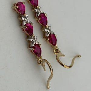 May include: A pair of gold-coloured drop earrings. Each earring features three teardrop-shaped, deep red gemstones. Small, silver-coloured accents are between the gemstones. The earrings have a curved hook design.