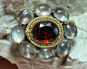 May include: A gold pendant with a large, dark red gemstone surrounded by eight smaller, clear gemstones.