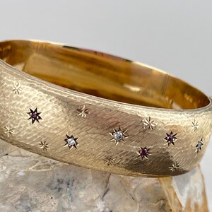 May include: A gold bangle bracelet with a hammered finish and a starburst design. The bracelet is set with small diamonds and rubies.