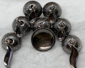 May include: A set of seven silver-toned metal ornaments, each with a decorative pattern and a dangling teardrop-shaped charm. The ornaments are arranged in a circular pattern, with a larger, flat ornament in the centre.