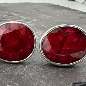Puede incluir: Un par de pendientes de botón de forma ovalada con piedras preciosas facetadas de color rojo intenso engastadas en biseles de metal plateado liso. Las piedras muestran inclusiones naturales y una superficie pulida que refleja la luz.