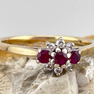 May include: A gold ring with a cluster of three deep red gemstones and smaller white stones. The ring has a simple gold band. The ring is set against a textured, neutral background.