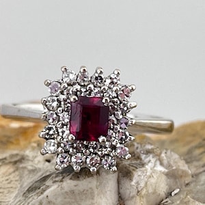 May include: A silver ring featuring a square ruby gemstone set within a halo of smaller diamonds.
