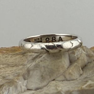 May include: A silver ring with a textured surface, resting on a light-colored stone. The ring has the markings "925 S" and "28 A" engraved on its surface. The ring is a band style, likely for everyday wear.