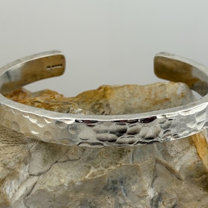 Könnte beinhalten: Ein silbernes Armreif-Armband mit gehämmertem Muster. Das offene Armband wird auf einer hellbraunen und beigefarbenen Steinoberfläche präsentiert. Das Armband hat ein schlichtes, elegantes Design.