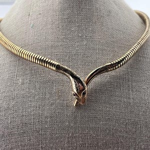 Może przedstawiać: Złoty naszyjnik w kształcie węża ze zwiniętym korpusem i szczegółową głową węża. Naszyjnik w stylu choker ma zapięcie w kształcie głowy węża. Oczy węża to małe, ciemne kamienie.