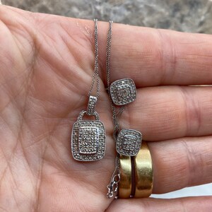 May include: A silver necklace with a square pendant and matching square stud earrings, both encrusted with small, sparkling stones. The jewelry is displayed on a hand, with two gold rings visible.