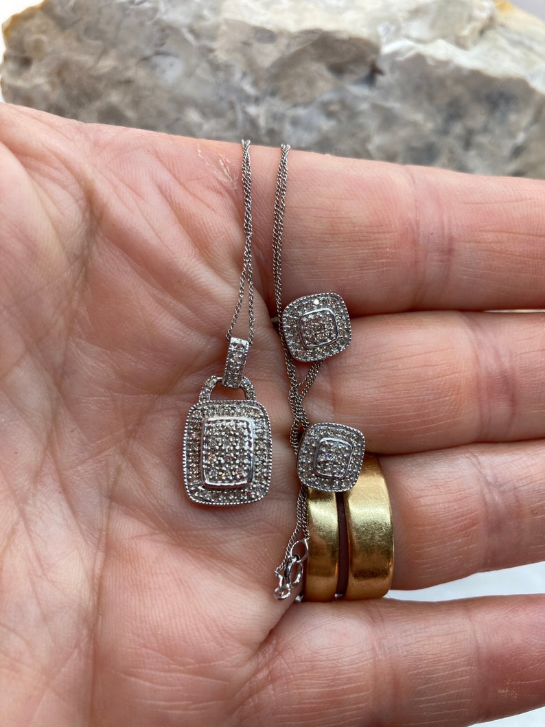 May include: A silver necklace with a square pendant and matching square stud earrings, both encrusted with small, sparkling stones. The jewelry is displayed on a hand, with two gold rings visible.