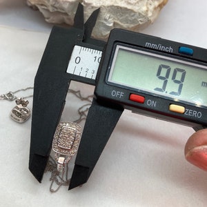 May include: A silver pendant necklace with a rectangular pendant, measuring 9.9 mm. The pendant is encrusted with small, clear stones. A digital caliper displays the measurement. The necklace chain is delicate.