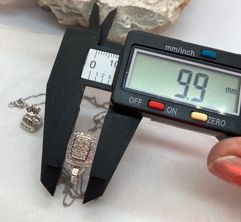 May include: A silver pendant necklace with a rectangular pendant, measuring 9.9 mm. The pendant is encrusted with small, clear stones. A digital caliper displays the measurement. The necklace chain is delicate.