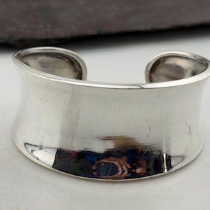 May include: A wide, polished silver cuff bracelet. The bracelet has a curved, open design with a smooth, reflective surface. The silver reflects the surrounding environment, creating a bright, metallic appearance. The cuff is a bold, statement piece of jewelry.