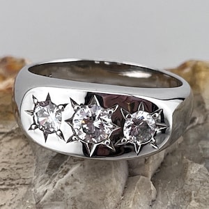 May include: A polished silver ring with three clear, starburst-shaped gemstones. The ring has a smooth, rounded band and the gemstones are set in a row across the top. The ring is displayed on a textured, light-coloured surface.