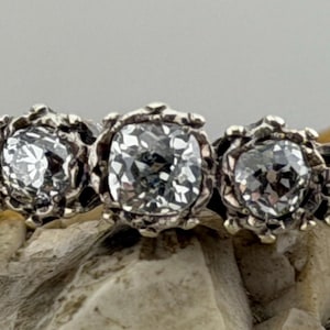 Könnte beinhalten: Ein Silberring mit fünf klaren, rund geschliffenen Edelsteinen. Die Edelsteine sind in kunstvollen, floral inspirierten Fassungen gefasst. Der Ring wird auf einer strukturierten, hellbraunen Oberfläche präsentiert.