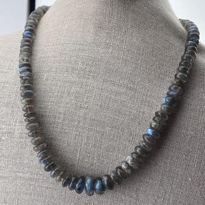 May include: A necklace made of dark grey labradorite beads with iridescent blue flashes. The beads are oval shaped and strung on a silver chain.