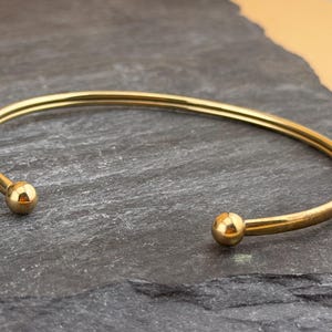 Könnte beinhalten: Ein goldfarbenes Armband aus Metall mit glatter, polierter Oberfläche und einem schlichten, offenen Design. Das Armband ist kreisförmig und endet an beiden Seiten mit einem kleinen, kugelförmigen Akzent, was für ein klares und einheitliches Aussehen sorgt.