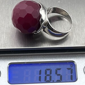 Op de afbeelding: Een zilveren ring met een grote, gefacetteerde, donkerrode edelsteen. De ring ligt op een digitale weegschaal die 18,57 gram aangeeft.