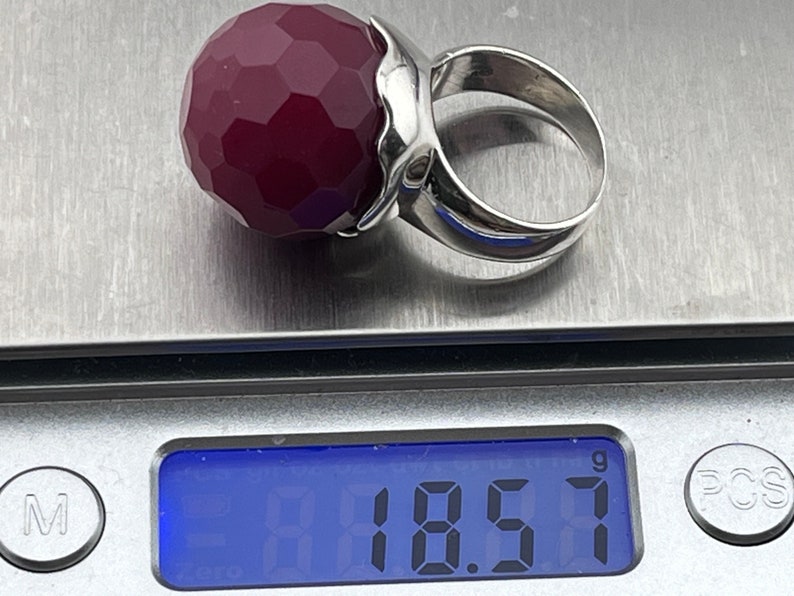 Op de afbeelding: Een zilveren ring met een grote, gefacetteerde, donkerrode edelsteen. De ring ligt op een digitale weegschaal die 18,57 gram aangeeft.