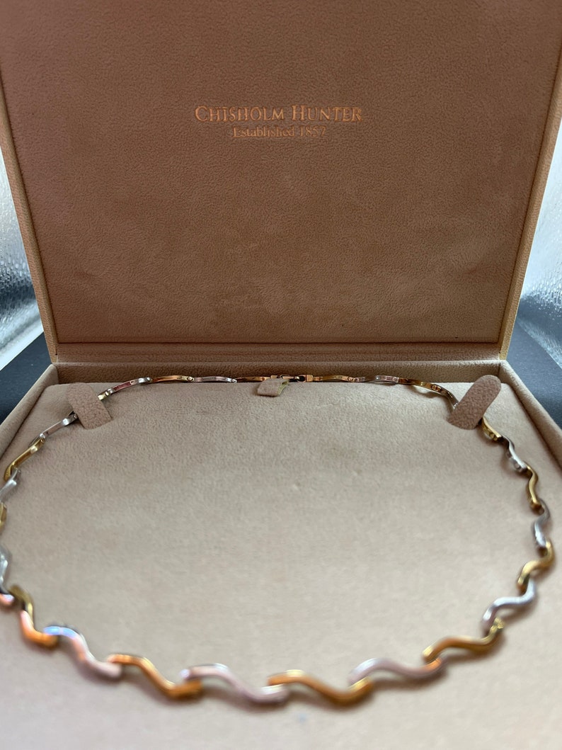 Op de afbeelding: Een gouden, zilveren en ros&eacute;gouden ketting met een golvend ontwerp. De ketting zit in een doos met de tekst "Chisholm Hunter Established 1857".