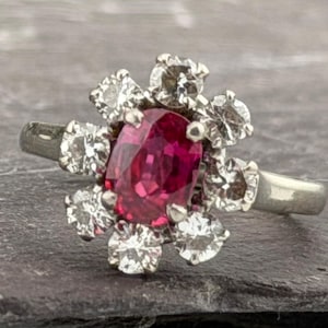 Puede incluir: Un anillo de tono plateado con una piedra preciosa central de corte ovalado en color rosa intenso, rodeada por un grupo de ocho piedras claras de corte redondo dispuestas en forma floral sobre una banda de metal pulido.