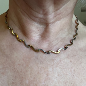 Op de afbeelding: Een zilveren en gouden ketting met een golvend ontwerp. De ketting is gemaakt van gekoppelde metalen stukken die afwisselend zilver en goud zijn.