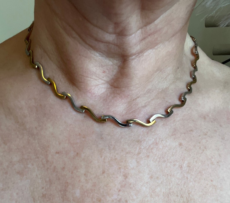 Op de afbeelding: Een zilveren en gouden ketting met een golvend ontwerp. De ketting is gemaakt van gekoppelde metalen stukken die afwisselend zilver en goud zijn.