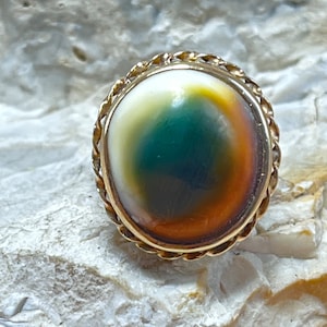 May include: A gold ring with a large oval-shaped gemstone. The gemstone is a mix of green, brown, and white colors.