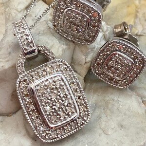 May include: A silver pendant necklace and matching square stud earrings. The pendant is a rectangular shape with a smaller rectangle in the center, all encrusted with small, sparkling stones. The earrings have a similar design. The jewelry set is displayed on a light-colored, textured surface.