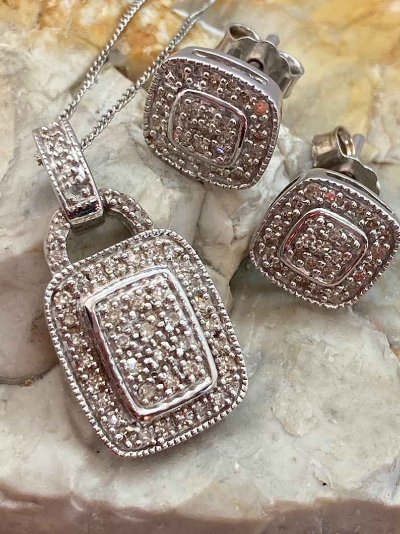May include: A silver pendant necklace and matching square stud earrings. The pendant is a rectangular shape with a smaller rectangle in the center, all encrusted with small, sparkling stones. The earrings have a similar design. The jewelry set is displayed on a light-colored, textured surface.