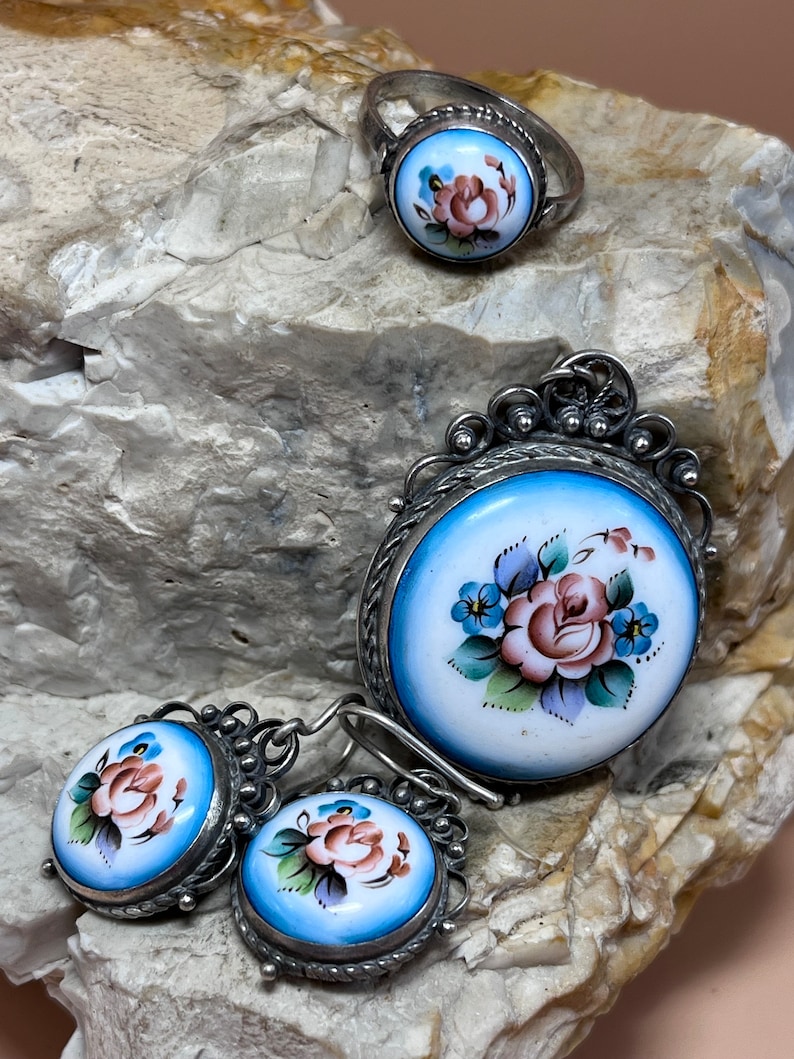 Peut inclure: Un ensemble de bijoux en argent avec &eacute;mail bleu et blanc orn&eacute; de motifs floraux peints &agrave; la main. L'ensemble comprend une bague, un pendentif et une paire de boucles d'oreilles.