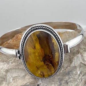 May include: A silver bangle bracelet with a large oval amber stone set in a silver bezel. The stone has a brown and yellow color and a unique pattern.
