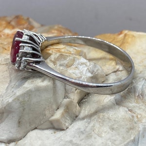 May include: A silver ring with a large, oval-shaped ruby gemstone surrounded by smaller diamonds. The ring is set on a white background.