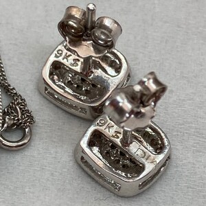 May include: Close-up of a pair of square-shaped stud earrings with a cluster of small stones. The earrings are made of silver-colored metal and have a post and backing. The earrings are marked with "9KS" and "DIA".
