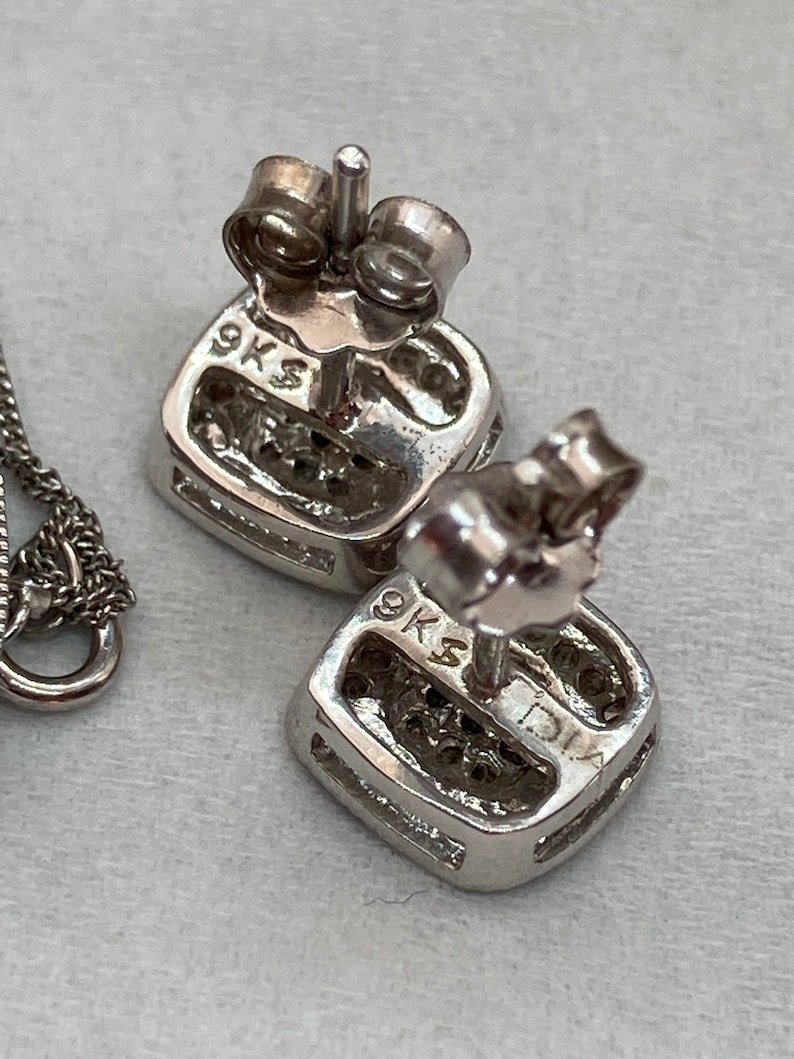 May include: Close-up of a pair of square-shaped stud earrings with a cluster of small stones. The earrings are made of silver-colored metal and have a post and backing. The earrings are marked with "9KS" and "DIA".