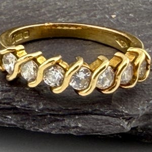 Op de afbeelding: Een gouden ring met een doorlopende rij van zeven ronde, heldere stenen gezet in een herhalend S-vormig metalen ontwerp. De binnenzijde van de gouden band toont zichtbare keurmerken en stempels die de zuiverheid van het metaal aangeven.