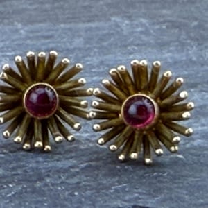 May include: A pair of stud earrings featuring a circular center set with a smooth, deep red gemstone. Radiating outward from the center are numerous thin, textured gold-toned metal spikes, each tipped with a small, rounded bead, creating a sunburst design.