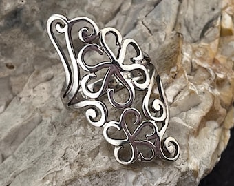 May include: A silver-colored ring with an openwork design featuring swirling patterns and floral shapes. The ring is set against a textured, light-colored stone surface, showcasing its intricate details.
