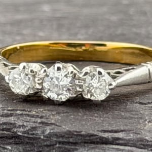 May include: A three-stone ring featuring a yellow gold band and a white metal setting. The ring showcases three round, clear, faceted stones arranged horizontally across the center, with intricate metalwork detailing along the shoulders.