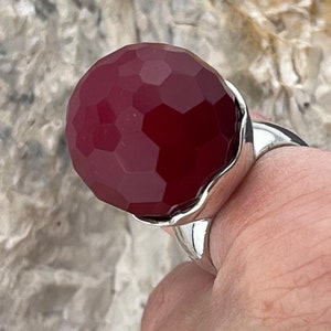 Op de afbeelding: Een zilveren ring met een grote, gefacetteerde, donkerrode edelsteen. De edelsteen is rond en heeft een glad, gepolijst oppervlak.