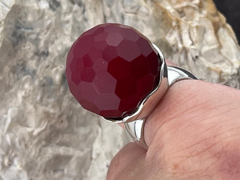 Op de afbeelding: Een zilveren ring met een grote, gefacetteerde, donkerrode edelsteen. De edelsteen is rond en heeft een glad, gepolijst oppervlak.