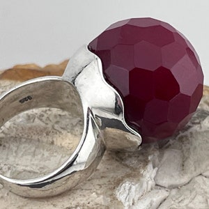 Op de afbeelding: Een zilveren ring met een grote, gefacetteerde, bordeauxrode edelsteen. De ring heeft een uniek, golvend ontwerp.