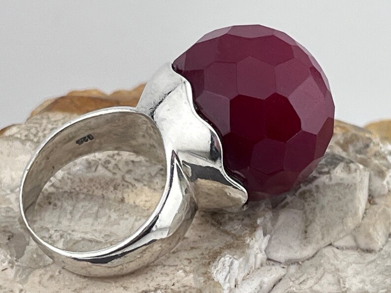 Op de afbeelding: Een zilveren ring met een grote, gefacetteerde, bordeauxrode edelsteen. De ring heeft een uniek, golvend ontwerp.