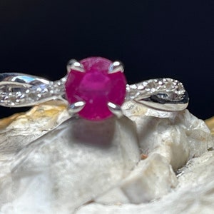 Può includere: Un anello d'argento con una pietra preziosa di rubino rosa e piccoli diamanti trasparenti sulla fascia.