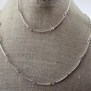 May include: A silver necklace and bracelet set. The jewellery features curved, slender bars connected by small circular links. The necklace and bracelet are displayed on a neutral fabric background, showcasing the minimalist design.