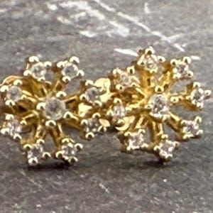 May include: A pair of gold-colored stud earrings. Each earring features a snowflake design with multiple clear, round gemstones. The earrings are set against a dark, textured background.