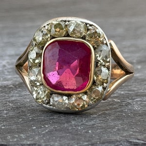 May include: A ring featuring a central cushion-cut deep pink gemstone set in a gold bezel. The centre stone is surrounded by a halo of twelve round-cut, light-coloured, faceted stones set in a silver-toned frame, mounted on a gold band.