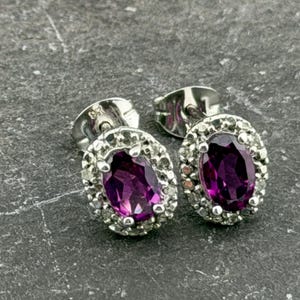May include: Silver stud earrings featuring oval, deep purple gemstones encircled by a halo of smaller, clear stones. The earrings are displayed on a textured, grey surface.