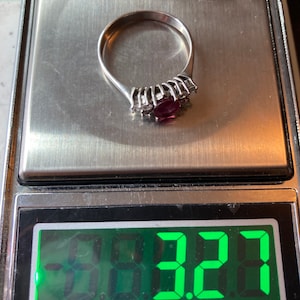 May include: A silver ring with a large, dark red gemstone surrounded by smaller clear gemstones. The ring is on a digital scale that reads 3.22 grams.